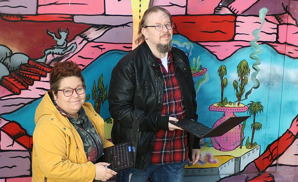 Piia Niilola ja Janne Haikka läppäreineen värikkään seinämaalauksen edessä.