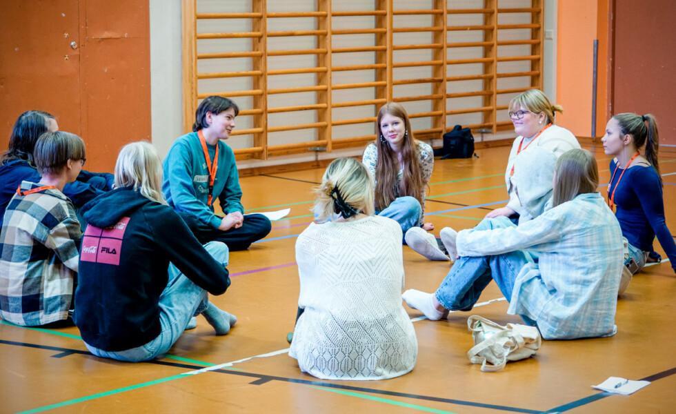 Nuoria istumassa salin lattialla ja keskustelemassa. Lattialla on kyniä ja lehtiöitä.