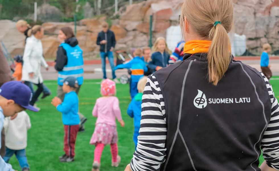 Joukko lapsia ja aikuisia kokoontuneella vihreälle nurmelle. Etualalla selkä kohti kameraa seisoo nainen, jonka liivin selkäpuolella lukee teksti "Suomen latu".