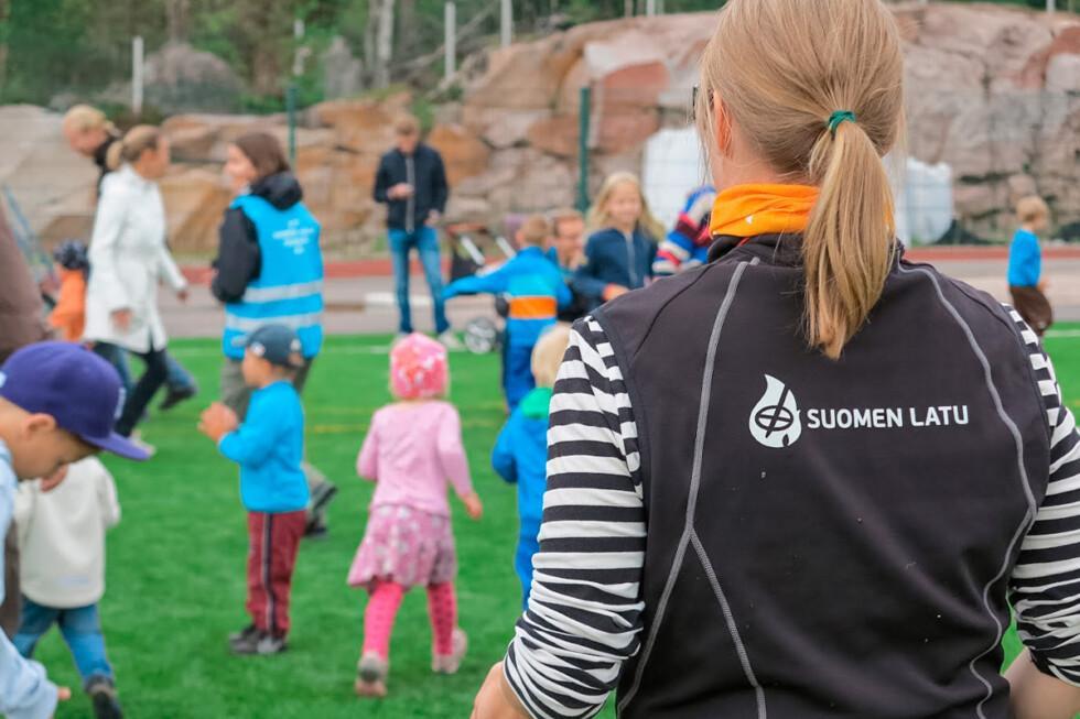 Joukko lapsia ja aikuisia kokoontuneella vihreälle nurmelle. Etualalla selkä kohti kameraa seisoo nainen, jonka liivin selkäpuolella lukee teksti "Suomen latu".