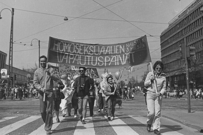 Mustavalkoinen valokuva kadulla marssivista ihmisistä. Etualalla marssivat mies ja nainen kantavat lippua, jossa lukee teksti "homoseksuaalien naisten ja miesten vapautuspäivät".