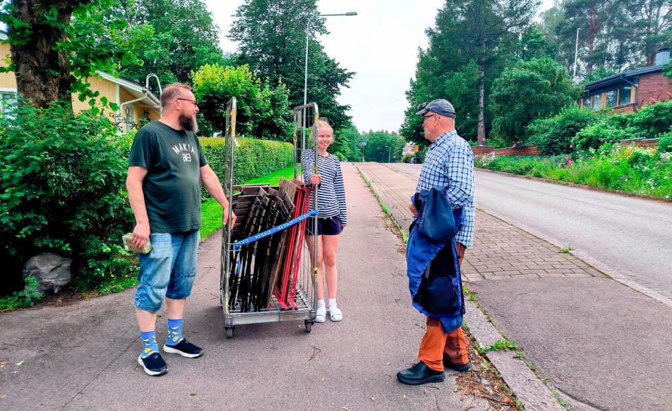 Parrakas mies ja vaaleahiuksinen tyttö pitävät jalkakäytävällä kiinni rullakosta, johon on koottu puutavaraa. Mies ja tyttö hymyilevät, ja heidän seurassaan on käsissään takkia pitelevä mies.