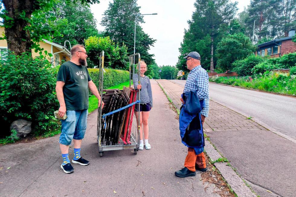 Parrakas mies ja vaaleahiuksinen tyttö pitävät jalkakäytävällä kiinni rullakosta, johon on koottu puutavaraa. Mies ja tyttö hymyilevät, ja heidän seurassaan on käsissään takkia pitelevä mies.