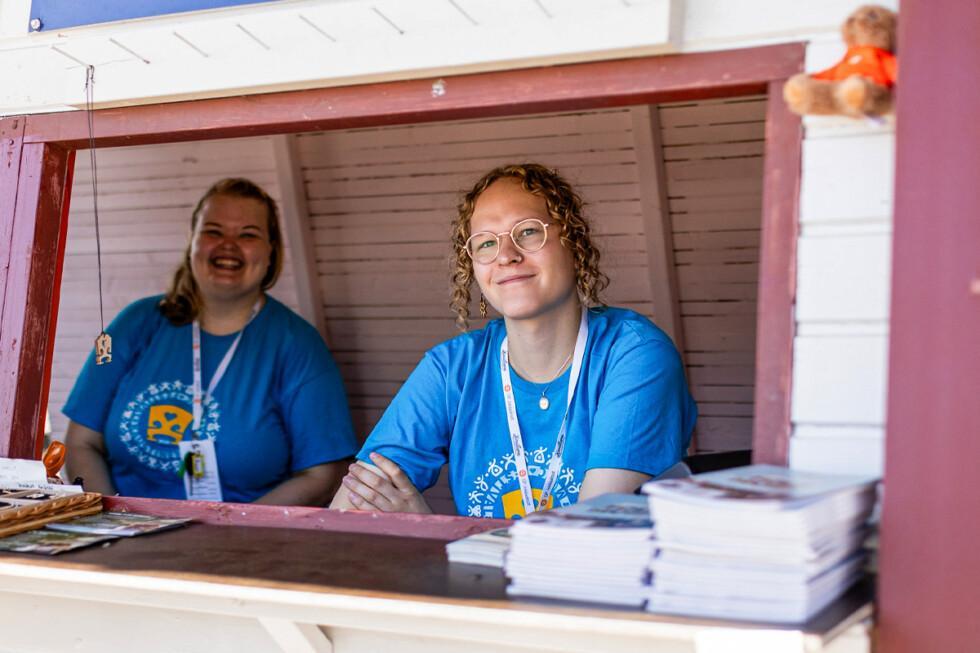 Kaksi talkoolaista katsoo hymyillen kameraan myyntikojusta käsin. Molemmilla on yllään sininen paita ja kaulanauha.