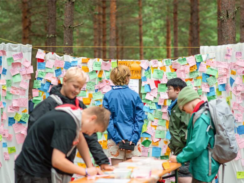 Nuoria kirjoittamassa post-it -lapuille viestejä. Taustalla suuri kangas, johon on kirjoitettu useita viestejä.